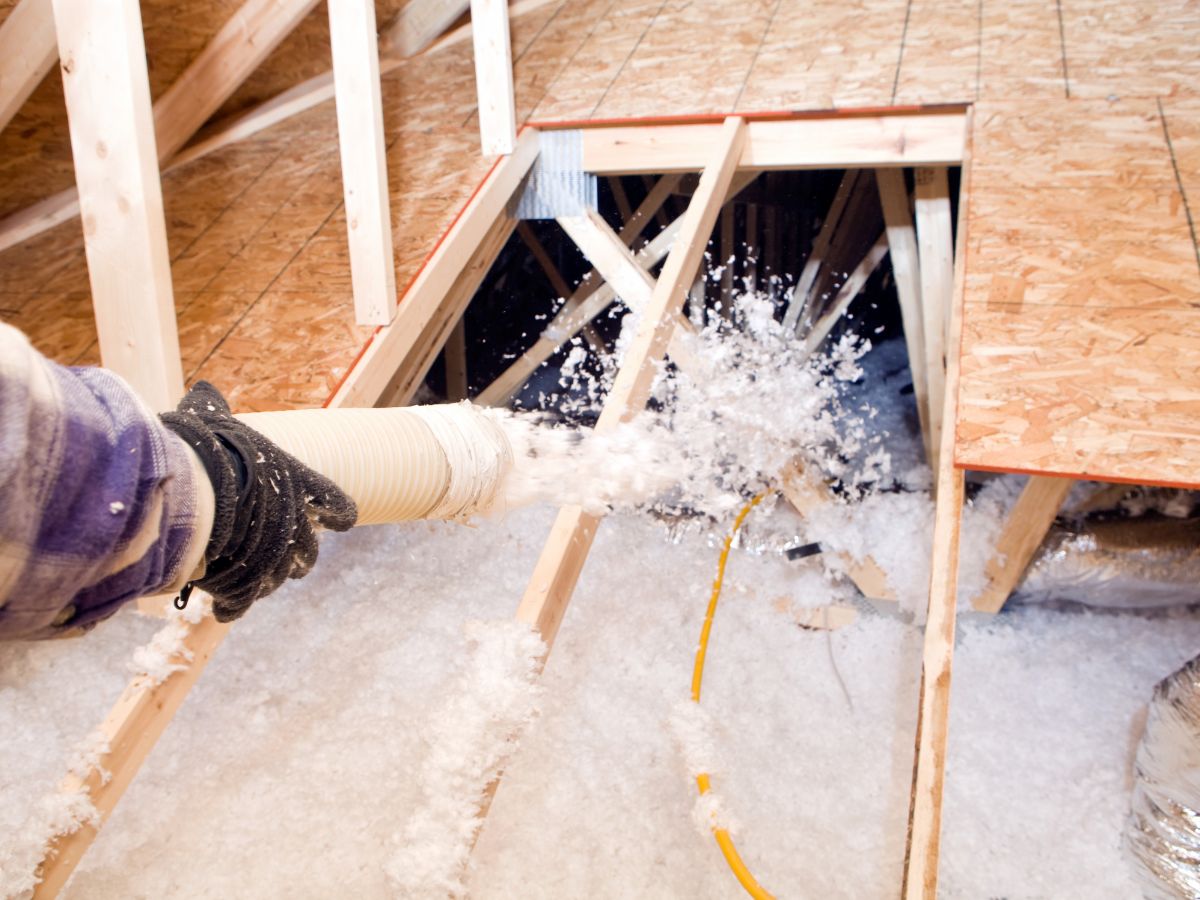 A gloved hand operates a hose, spraying insulation material into an attic space, with wooden beams and flooring visible.