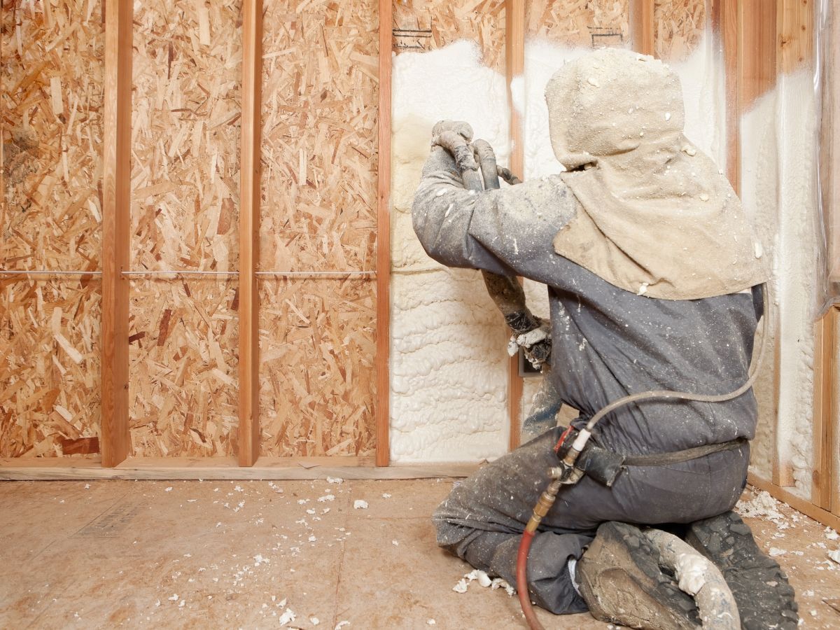 A worker in protective gear kneels to apply insulation to a wooden wall structure in a construction setting.