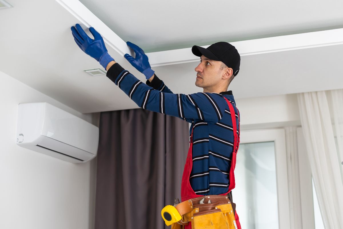 A worker in a striped shirt and red overalls adjusts a ceiling panel, wearing blue gloves and a tool belt in a bright room.
