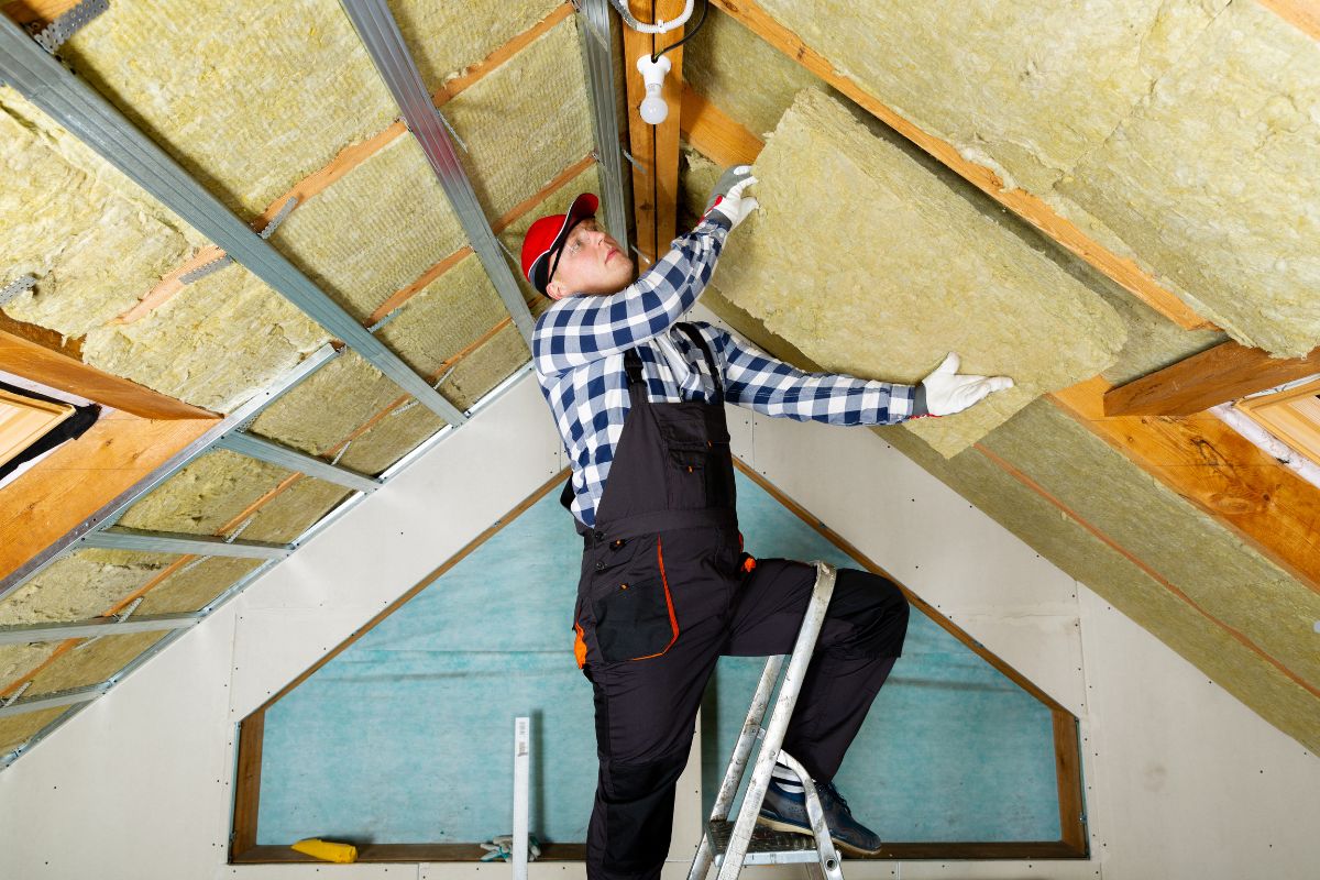 A worker on a ladder installs insulation material in an attic, surrounded by wooden beams and drywall.