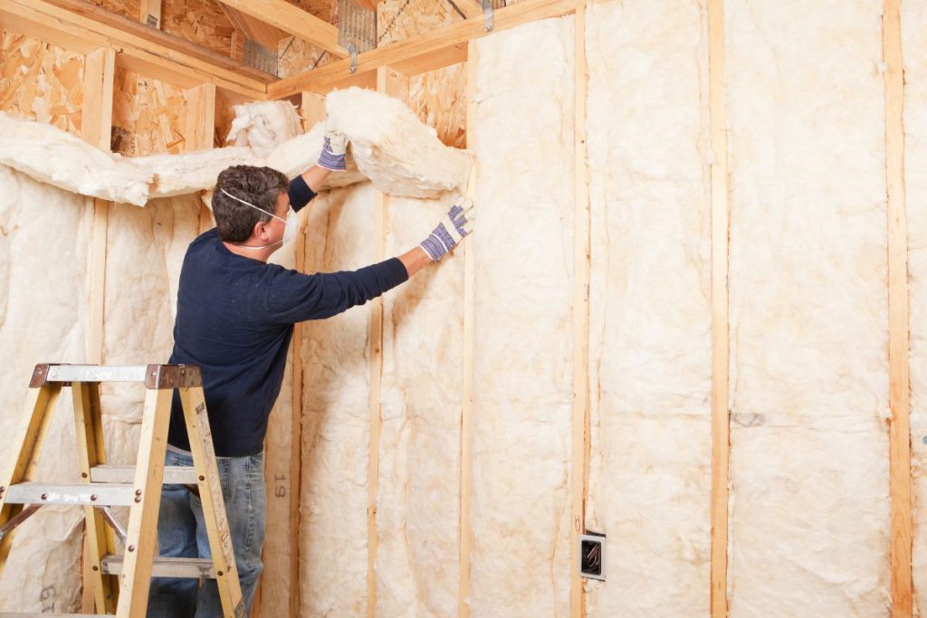 A person installs insulation material in a wooden framework, standing on a ladder with tools and gloves, focused on the task.