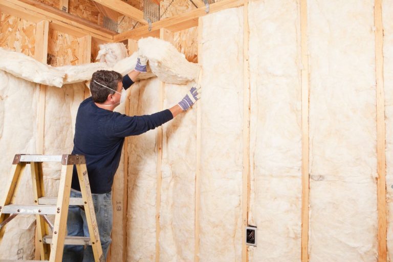 A person installs insulation material in a wooden framework, standing on a ladder with tools and gloves, focused on the task.